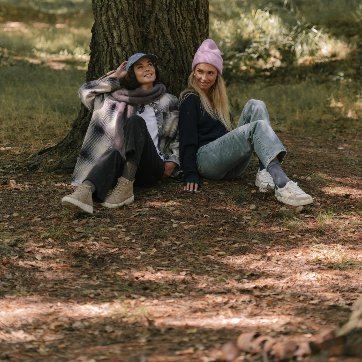 Two women sitting under a tree, wearing casual shoes and cozy fall outfits in a forest setting.