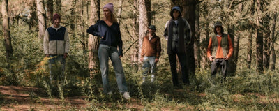 Group of people exploring a forest dressed in autumn attire, including hats and jackets, surrounded by trees and natural greenery.